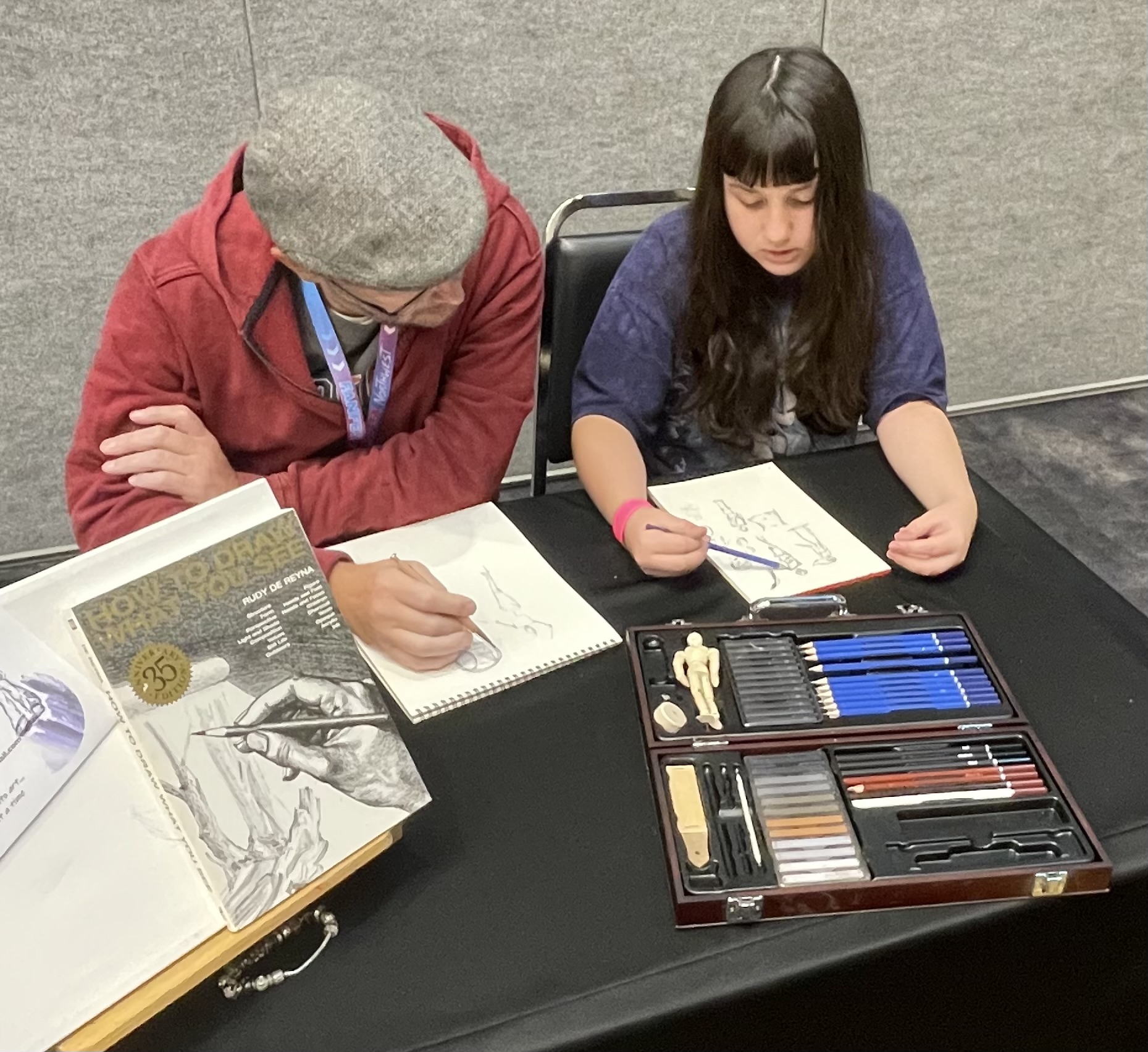 a girl working on a sketchbook with an art instructor drawaing at a talbe.